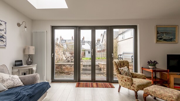 A modern living room with a sofa, armchair, and ottoman, a TV stand, and a large sliding glass door leading to an outdoor area. - Devon Cottage | Elie (Elie)