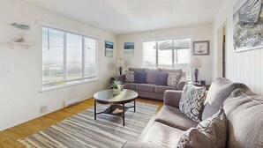 A living room with a sofa, a coffee table, and a rug. There are two windows with blinds, a lamp, and a picture on the wall. - Bodega Bay 2BR ocean-view cottage with hot tub — dog friendly (Bodega Bay)