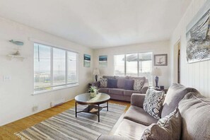 A living room with a sofa, a coffee table, and a rug. There are two windows with blinds, a lamp, and a picture on the wall.