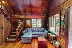 A wooden cabin interior with a sectional sofa, a staircase, and large windows. - Cazadero 1BR A-frame cabin with hot tub for two — dog friendly (Cazadero)