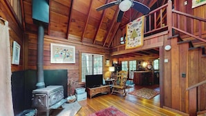 A wooden cabin interior with a wood stove, a television, a rocking chair, and a staircase leading to an upper level. - Cazadero 1BR A-frame cabin with hot tub for two — dog friendly (Cazadero)