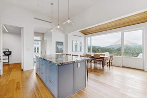 A modern kitchen with a large island, wooden flooring, pendant lighting, and a dining area with a view.