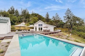 A swimming pool with lounge chairs and umbrellas, a small white building, and a forested hillside.
