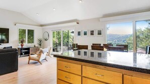 Uma sala de estar ampla com uma grande janela, cozinha com bancada de granito e área de jantar com cadeiras.
