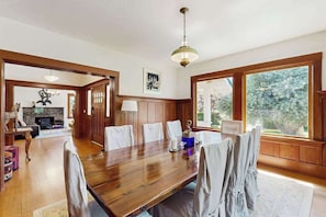 Ein Esszimmer mit Holztisch und -stühlen, einer Chandelüster und großen Fenstern mit Blick auf Bäume.