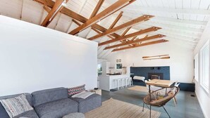 Ein modernes Interieur mit hohen Decken, freiliegenden Holzbalken, einem grauen Sofa, einem gewebten Teppich und einer Essecke mit Tisch und Stühlen.