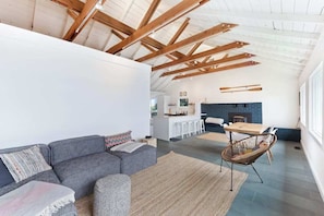 A modern interior with a high ceiling, exposed wooden beams, a grey sofa, a woven rug, and a dining area with a table and chairs. - Bodega Bay 2BR bay-view bungalow with private sauna — dogs welcome (Bodega Bay)