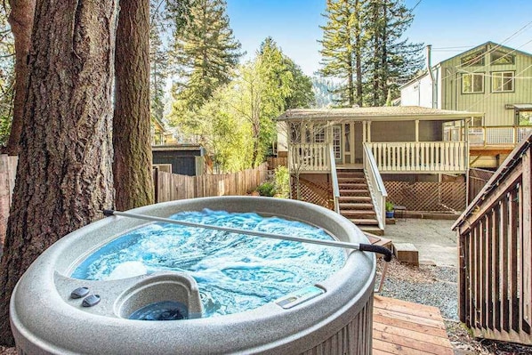 A hot tub in a backyard with a wooden deck, a house, and trees.