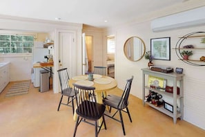 A dining area with a round table, four chairs, a small kitchen area with a refrigerator, and a wall-mounted shelf with various items.