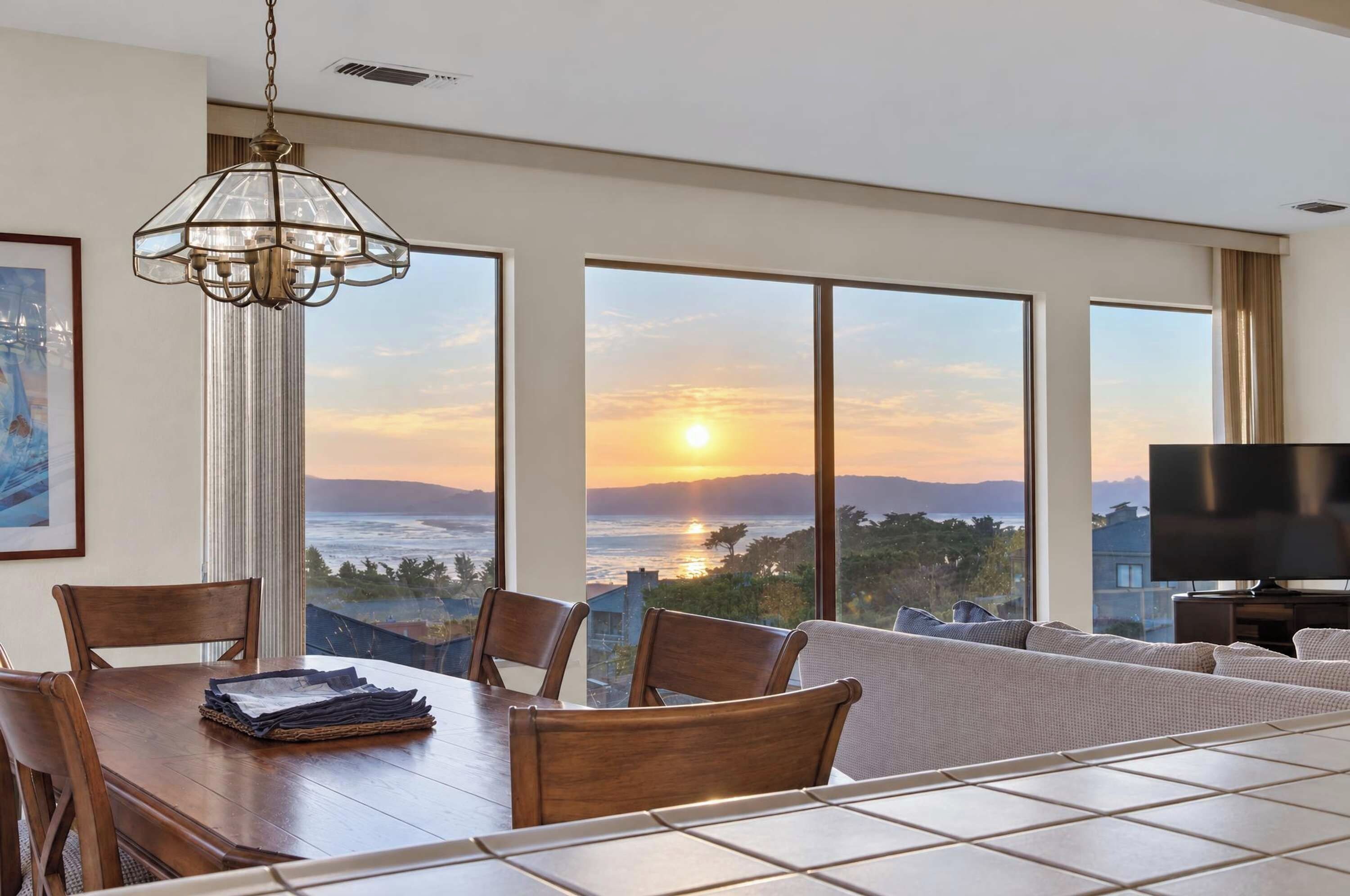 A modern living room with a dining area, large windows with a sunset view, and a flat-screen TV.