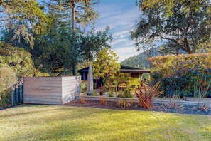 Uma área de estar moderna ao ar livre, com um deck de madeira, espaço coberto para sentar e um jardim com diversas plantas e árvores.