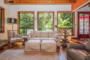 A cozy living room with a sofa, ottoman, and chair, large windows with a view of greenery, and a wooden ceiling. - Cazadero 4BR mountain cabin with hot tub in the redwoods — dog friendly (Cazadero)