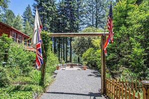 A pathway leading to a wooden structure with flags, surrounded by dense forest. - Cazadero 4BR mountain cabin with hot tub in the redwoods — dog friendly (Cazadero)