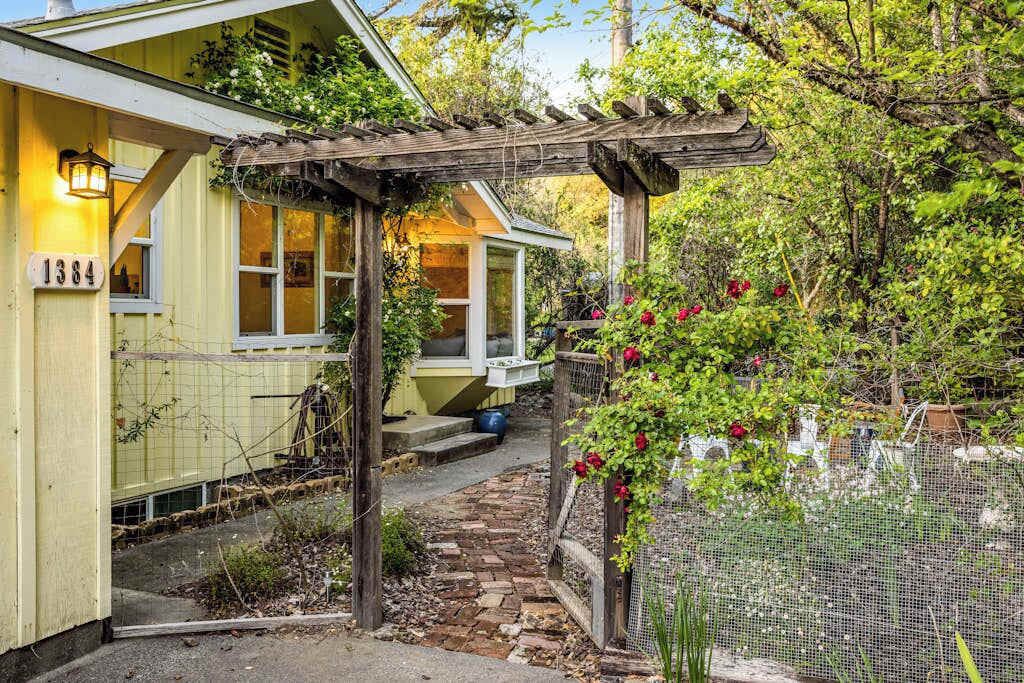 Un bâtiment jaune, avec le numéro « 1384 » inscrit sur le côté, une pergola en bois et un jardin parsemé de fleurs rouges.