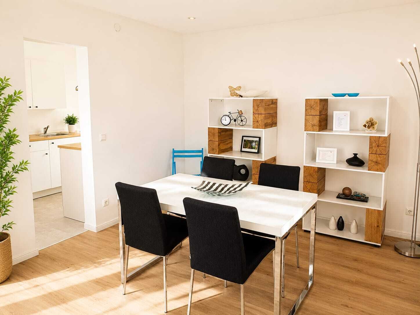 A dining area with a white table, black chairs, and a wooden floor. There is a kitchen visible in the background.