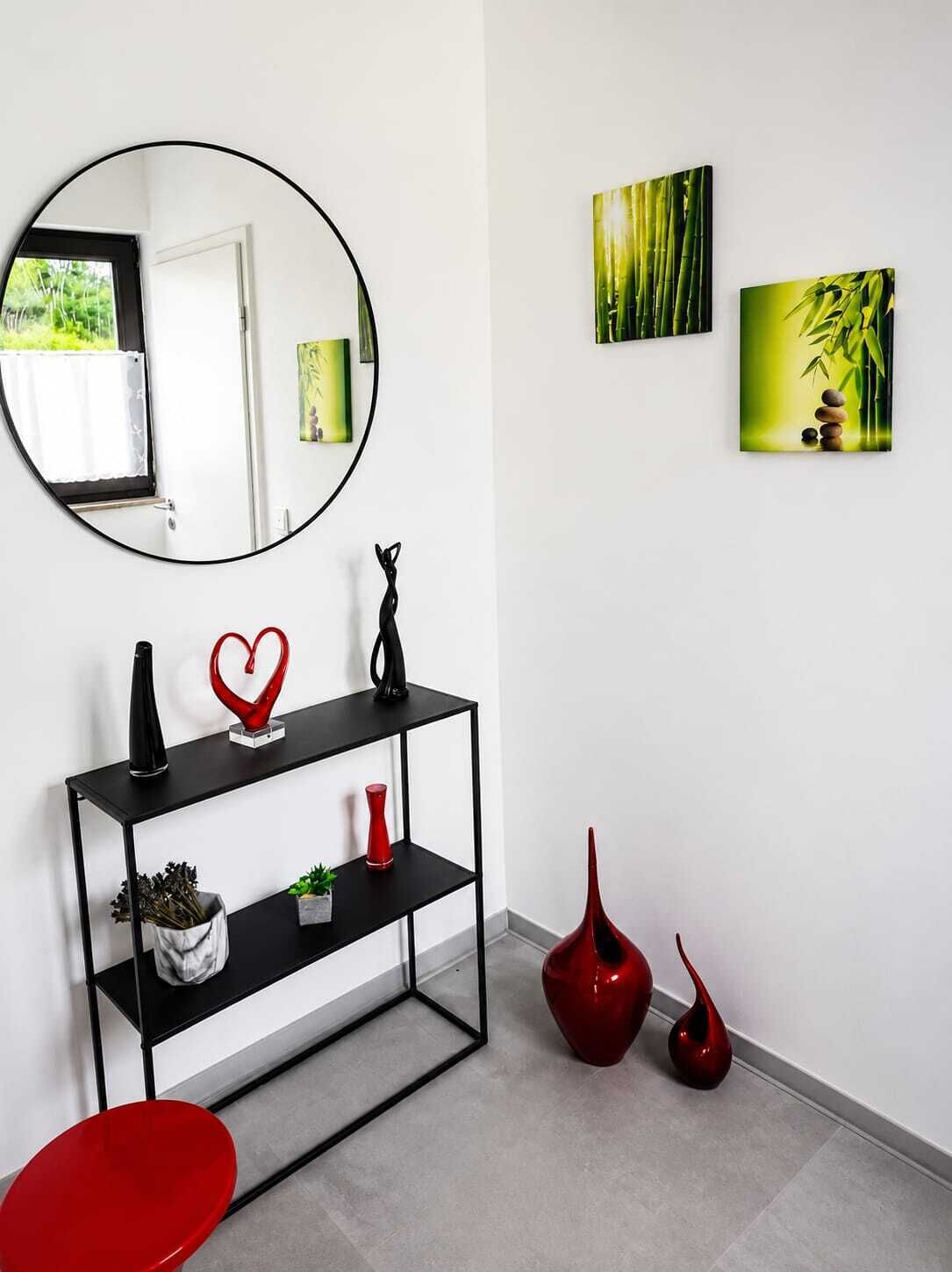 Ein modernes Interieur mit einem runden Spiegel, einem schwarzen Konsolentisch mit Dekorationselementen, roten Vasen und zwei gerahmten Kunstwerken an der Wand.