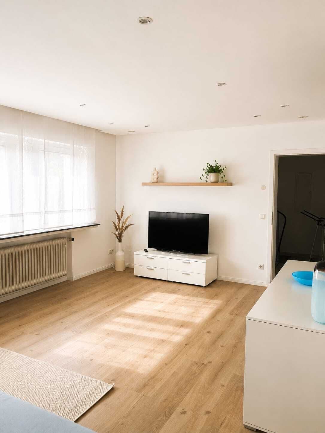 Una sala de estar moderna con televisor de pantalla plana, muebles blancos, piso de madera y ventana con cortinas traslúcidas.