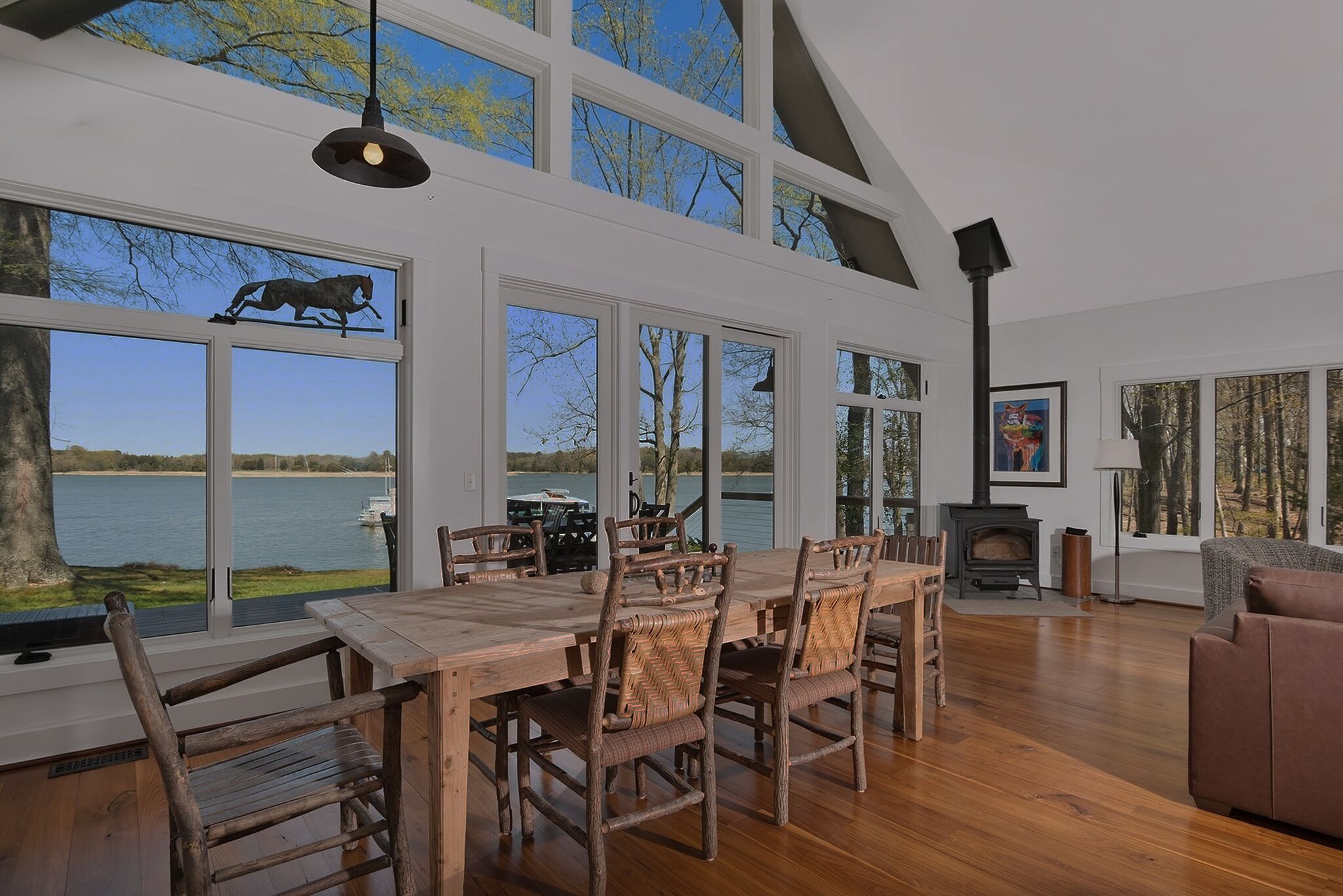 Une pièce spacieuse avec une grande table à manger en bois, des chaises en osier, une cheminée et de grandes fenêtres donnant sur un lac.