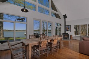 Ein geräumiges Zimmer mit einem großen Holzesstisch, Korbstühlen, einem Kamin und großen Fenstern mit Blick auf einen See.