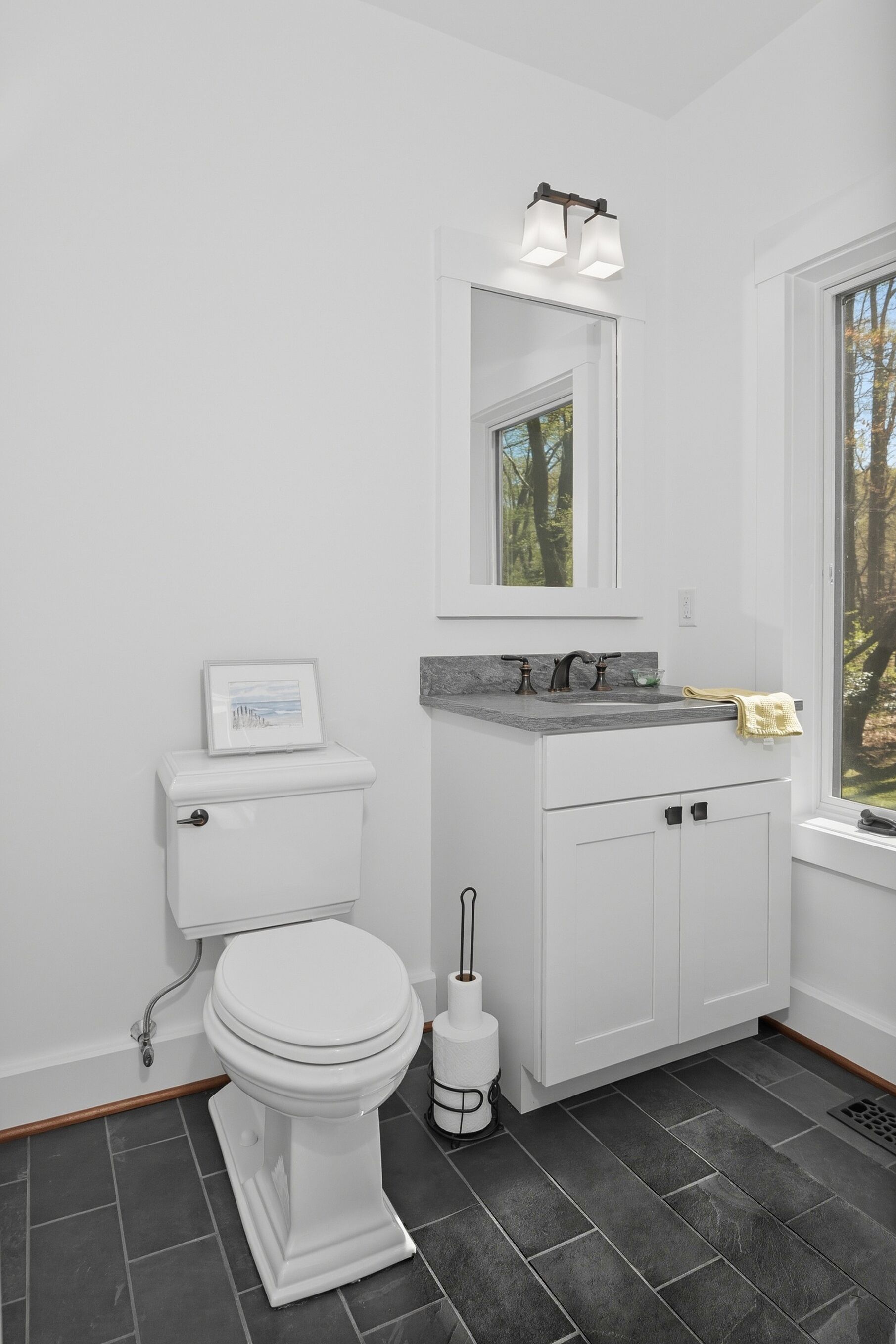 Une salle de bain avec des toilettes blanches, un lavabo doté d’un plan de travail en marbre et une fenêtre donnant sur des arbres.