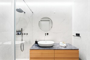 Un bagno moderno con cabina doccia in vetro, parete rivestita in marmo, mobile bagno in legno, specchio rotondo e lavandino bianco.