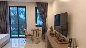 A modern hotel room with a bed, a desk with a flat-screen TV, a small table with a vase, chairs, and a view of palm trees through the window. - July Hotel Quang Binh (Dong Hoi)