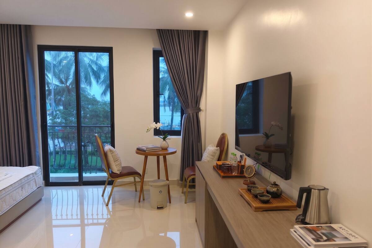 Habitación de hotel moderna con cama, escritorio con televisor de pantalla plana, una mesita con un jarrón, sillas y vista a palmeras por la ventana.