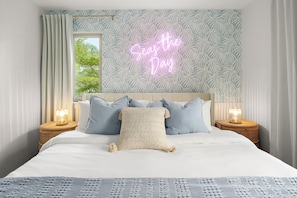 A neatly made bed with a 'Seas the Day' neon sign, two bedside lamps, and a window with curtains.