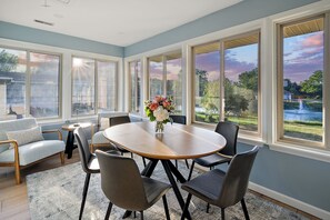 A dining area with a round table, six chairs, a vase of flowers, and large windows with a view of a garden and a lake. - 4BR Serene Waterfront Retreat, Lewes · 3 King Beds, Linens Included, Elevator (Lewes)