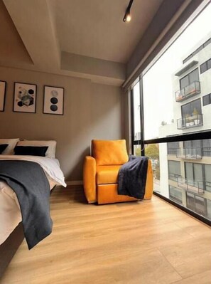 Ein modernes Hotelzimmer mit einem Bett, einem einzelnen orangefarbenen Sessel und großen Fenstern mit Blick auf das Außere des Gebäudes.