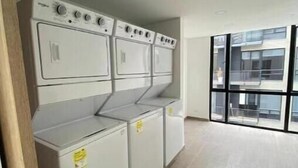Un cuarto de lavandería con lavadoras y secadoras apiladas, piso de baldosas y un ventanal con vista a un edificio.