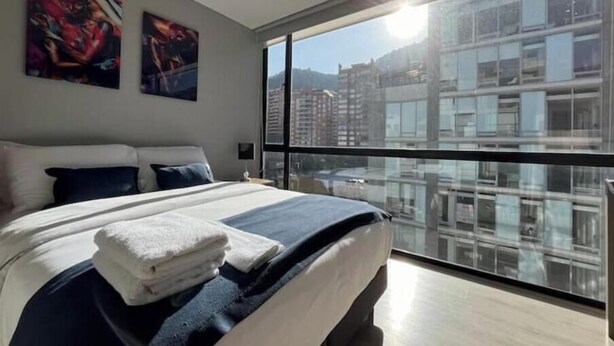 Un hotel moderno con una cama grande, dos cuadros en la pared y vistas a un paisaje urbano a través de un ventanal.