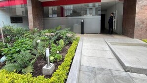 Una entrada moderna de edificio con mostrador de recepción, un jardín ajardinado con diversas plantas y un camino despejado hacia la entrada.