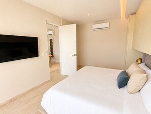 A hotel room with a bed, a TV mounted on the wall, an air conditioning unit, and a door leading to a bathroom. - Modern 2BR Condo - Rooftop Pool + Prime Location (Playa del Carmen)