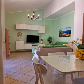 Ein Wohnzimmer mit Fernseher, Sofa, Esstisch, Stühlen und einer Kristallleuchter.