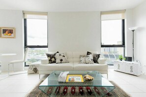 A modern living room with a white sofa, two large windows, a glass coffee table, and a city view. - Stylish2BD Penthouse Panoramic Views Nth Melbourne (North Melbourne)