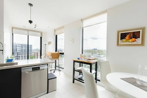 A modern kitchen with a dishwasher, a dining area with a table and chairs, and large windows offering a city view. - Stylish2BD Penthouse Panoramic Views Nth Melbourne (North Melbourne)