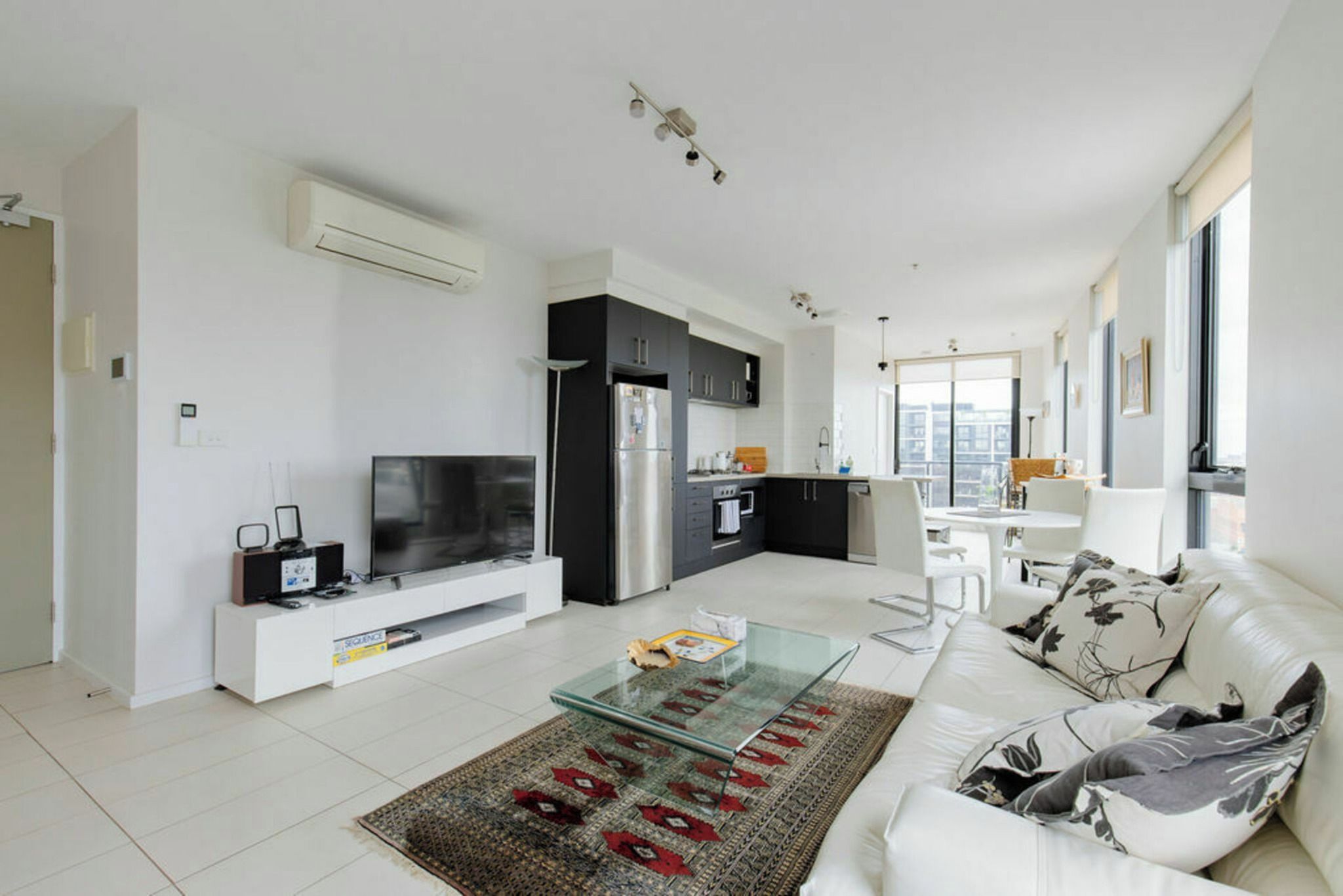 A modern living room with a glass coffee table, a sofa, a TV, and a kitchen area with a refrigerator and oven.
