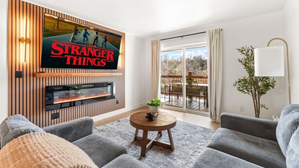 Een woonkamer met een flatscreen televisie bevestigd aan een houten paneel, een open haard, grijze banken, een ronde salontafel, en een schuifdeur van glas die leidt naar een buitenruimte.