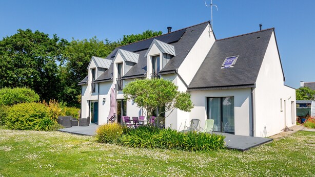 A modern two-story house with a sloped roof, large windows, and a small garden area with outdoor seating.