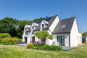Una casa moderna de dos pisos con techo inclinado, ventanas amplias y un pequeño jardín con mobiliario exterior.