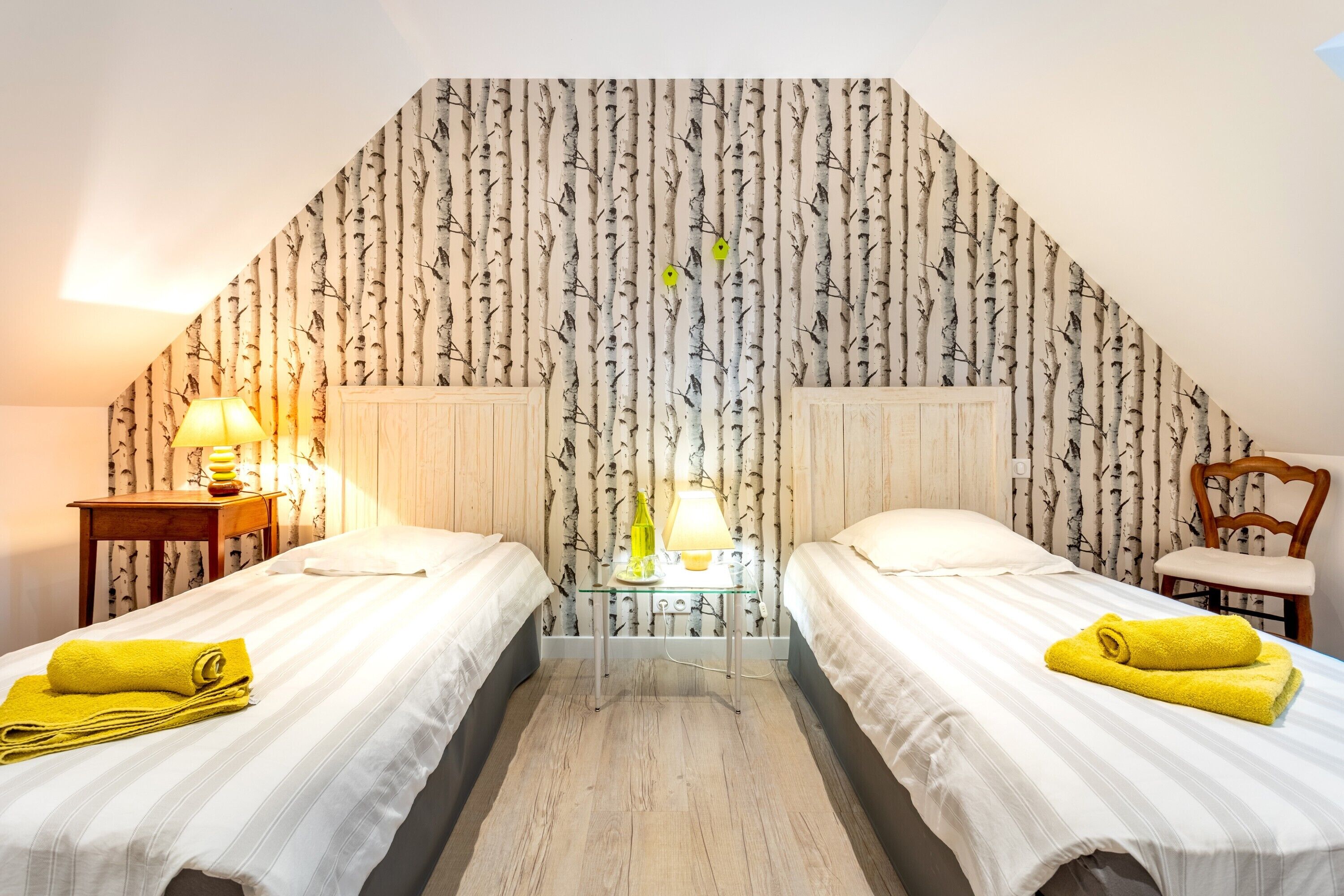 Une chambre avec deux lits, une table de chevet avec une lampe, une chaise et un mur orné d’un motif d’arbre.