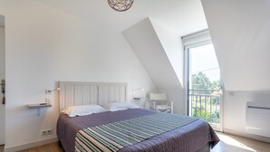Une chambre avec un plafond en pente, un lit recouvert d’une couvre-lit à rayures, une table de chevet avec une lampe, et une vue sur l’extérieur par une fenêtre.
