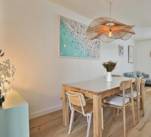 A dining area with a wooden table, white chairs, a hanging light fixture, and a wall-mounted painting.