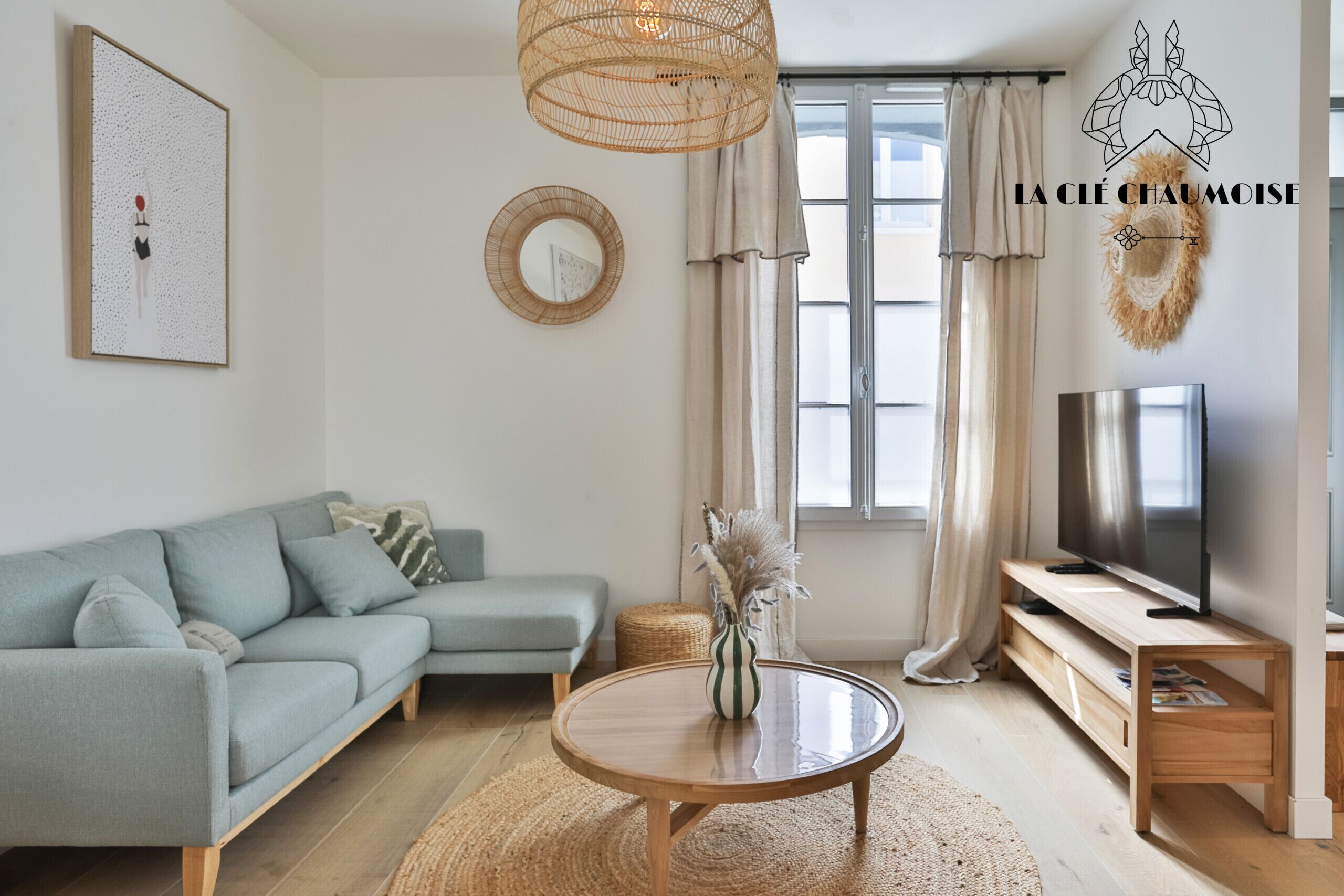 Un salon moderne avec un canapé, une table ronde en bois, un téléviseur et une grande fenêtre avec des rideaux.