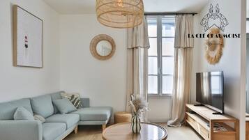 Un salon moderne avec un canapé, une table ronde en bois, un téléviseur et une grande fenêtre avec des rideaux.