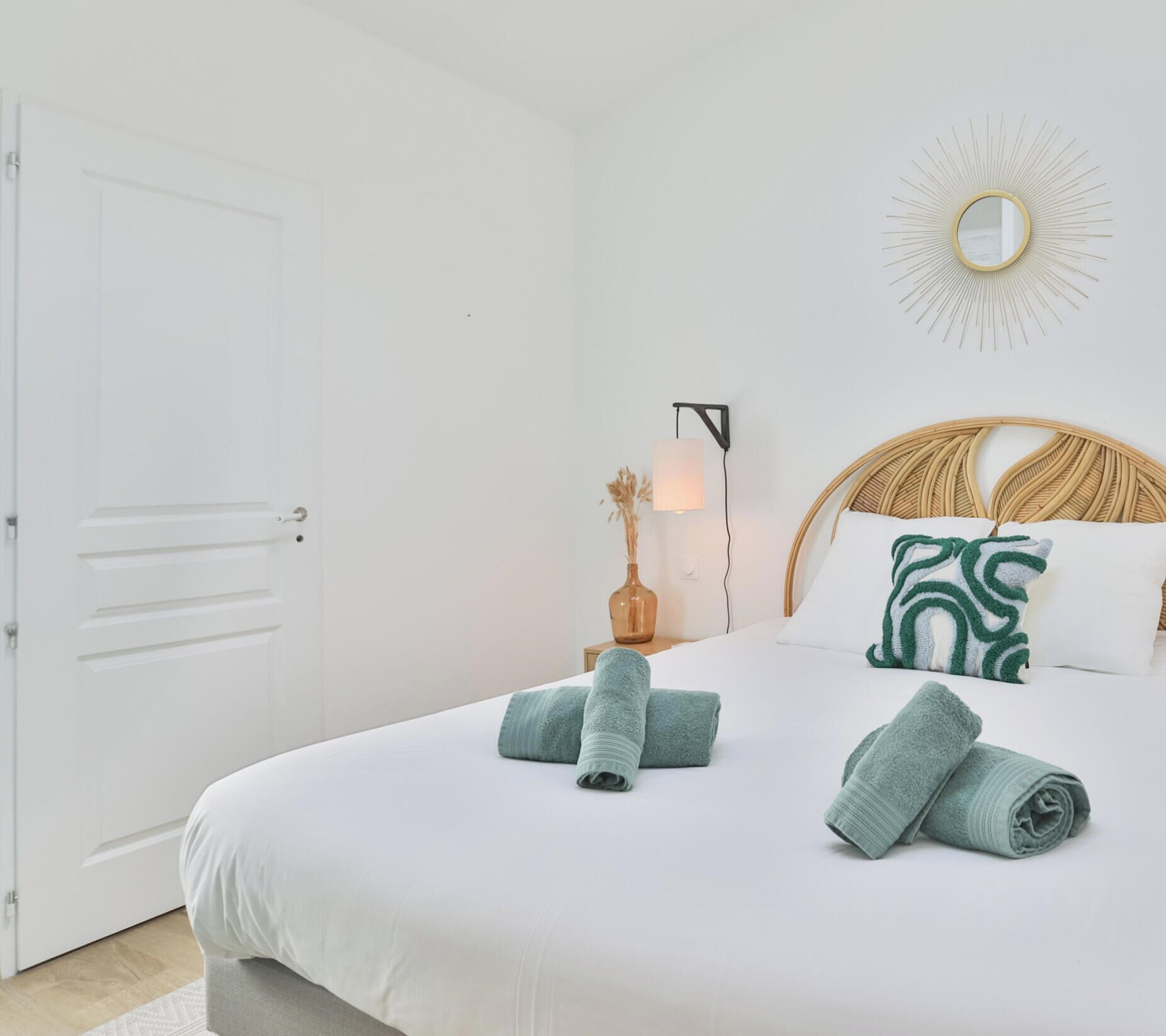 Un lit bien fait, agrémenté de serviettes pliées, un miroir fixé au mur et un luminaire décoratif en forme de soleil.