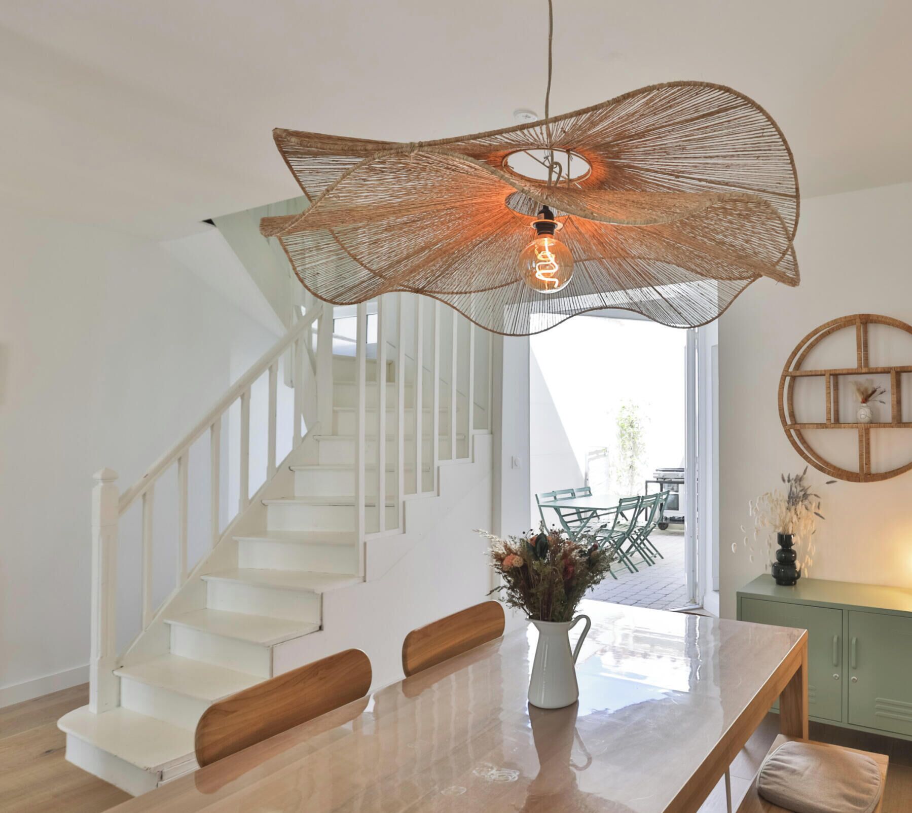 Un intérieur moderne avec un escalier, un coin repas agrémenté d’un vase de fleurs et un luminaire original en fibres tressées.