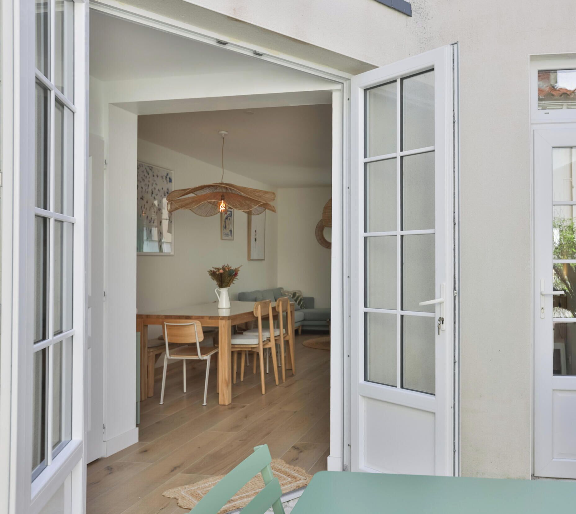 Une porte ouverte donne sur un espace repas où se trouvent une table et des chaises en bois, un luminaire suspendu et un vase rempli de fleurs.