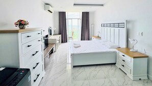 A modern hotel room with a bed, a desk, a dresser, and a view of the outdoors. - Căn Hộ View Biển (Phú Thủy)